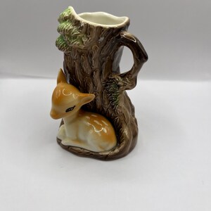 May include: A ceramic vase shaped like a tree trunk with a fawn nestled at the base. The vase is brown with green leaves and a white interior.
