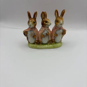 May include: A ceramic ornament depicting three rabbits in pink capes, standing on a green base. The rabbits have brown ears and are arranged side by side, with their arms around each other. A decorative item.
