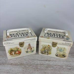 May include: Two vintage Peter Rabbit presentation boxes. Each box is off-white with a white handle and gold-coloured clasp. The boxes feature illustrations of Peter Rabbit and other characters from the Beatrix Potter stories. Text on the boxes reads "Peter Rabbit by Beatrix Potter."