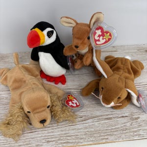 May include: Four Beanie Baby soft toys: a puffin, a kangaroo, a dog, and a rabbit. The puffin is black, white, red, and yellow. The kangaroo, dog, and rabbit are brown. Each toy has a heart-shaped tag.
