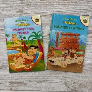 May include: Two hardback books featuring The Flintstones characters. The left book is titled Swimming Pool Pranks and shows Fred Flintstone lounging. The right book is titled Mistaken Identities and shows Fred and Barney in a stone car.