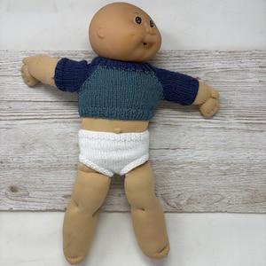 May include: A vintage doll with a tan complexion, freckles, and a smiling expression. The doll is wearing a two-tone blue knit sweater and white knit diaper. The doll's arms and legs are made of a soft, flesh-toned material.