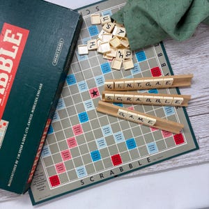 May include: A close-up of a Scrabble game board with tiles spelling out the words "V.I.N.T.G.A.E." and "S.C.R.A.B.B.L.E." on wooden racks. The tiles are arranged on a green and white game board with a red and blue grid.
