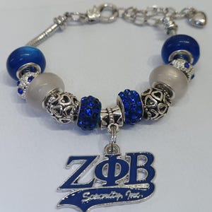 Puede incluir: Pulsera de abalorios plateada con cuentas azules y plateadas. La pulsera incluye un dije con las letras ZΦB en azul y las palabras "Sorority Inc." en blanco. Cierre de mosquetón.