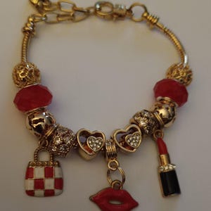 May include: Gold-toned charm bracelet with red and gold beads. Charms include a red and white checkered handbag, heart-shaped charms with rhinestones, a red lipstick, and red lips.