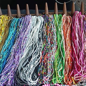 Puede incluir: Exhibición de collares de cuentas coloridas en varios tonos, incluyendo rojo, amarillo, azul, morado, blanco, verde, naranja, rosa y lavanda. Los collares cuelgan de clavijas de madera, mostrando la variedad de colores y estilos.