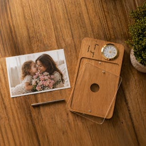 Op de afbeelding: Houten fotolijst met een kleine klok en de tekst "I love you mom". De lijst heeft een doorzichtige acryl cover en een cirkelvormige opening. Een foto van een moeder en kind ligt naast de lijst.