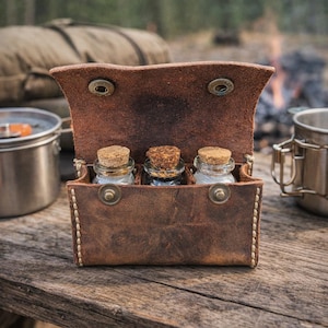 May include: A brown leather spice rack with three glass jars, each sealed with a cork stopper. The rack is open, revealing the jars, and is secured with metal snaps. The background shows camping gear and a campfire.