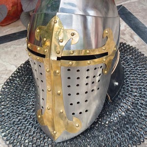 May include: A polished steel knight's helmet with brass detailing. The helmet has a cross design and ventilation holes. It sits on a chainmail coif, representing medieval armour.