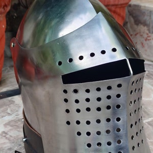 May include: A silver helmet with a perforated visor and a leather strap. The helmet has a rounded top and a pointed chin guard.