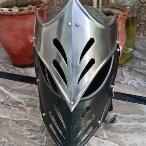 Pode incluir: Um capacete de estilo medieval com uma parte superior prateada polida e uma seção inferior de couro preto. O capacete apresenta recortes decorativos e rebites, sugerindo um tema histórico ou de fantasia.