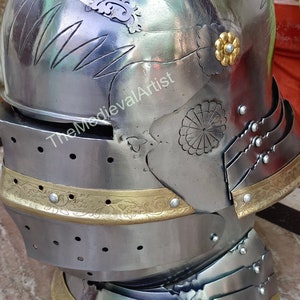 May include: A silver helmet with gold accents and intricate floral designs. The helmet has a visor and a neck guard. The helmet is on a marble surface. The text "The Medieval Artist" is visible on the helmet.