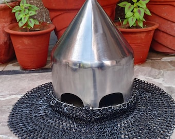 May include: A polished silver medieval helmet with a conical top, featuring eye openings and a chainmail neck guard. The helmet is displayed on a stone surface, with potted plants in the background.
