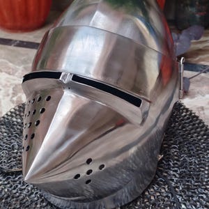 May include: A silver helmet with a pointed visor and a chainmail coif. The helmet has a series of small holes around the visor and on the sides.