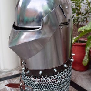 May include: A silver helmet with a chainmail coif and a hinged visor. The helmet has a rounded top and a pointed chin guard.