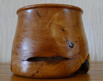 May include: A wooden vase with a natural, rustic finish. The vase has a wide mouth and a narrow base, and it is made from a single piece of wood. The wood has a rich brown color and a natural grain pattern.