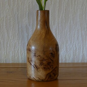 May include: A handcrafted wooden vase with a bottle-like shape. The vase is made of light-colored wood with dark, swirling grain patterns. It stands on a wooden surface and contains green stems.