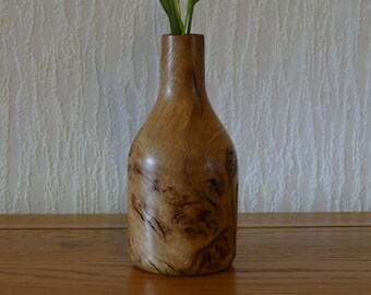 May include: A handcrafted wooden vase with a bottle-like shape. The vase is made of light-colored wood with dark, swirling grain patterns. It stands on a wooden surface and contains green stems.