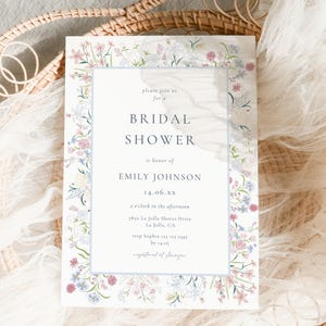 May include: A bridal shower invitation with a floral border, featuring the text "BRIDAL SHOWER" and details for Emily Johnson. The design includes a light blue frame and floral illustrations in pink, blue, and white.