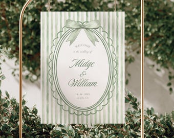 Puede incluir: Un letrero de bienvenida de boda rectangular colgado de un soporte de metal dorado. El letrero presenta un fondo de rayas verticales en verde salvia y blanco con un marco ovalado central, un lazo verde salvia y el texto: Welcome to the wedding of Midge & William.