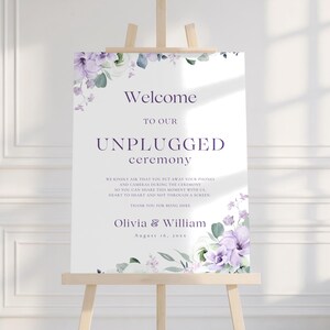Puede incluir: Un letrero blanco sobre un caballete de madera con las palabras "Welcome to our Unplugged Ceremony". Acentos florales en morado y verde enmarcan el texto. El letrero también incluye los nombres Olivia & William y la fecha 16 de agosto de 20xx.