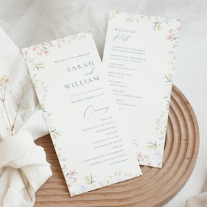 May include: Two wedding programs with floral borders. The programs display the names "SARAH and WILLIAM" and wedding details. The design includes a delicate floral border in pastel colours, with the text in a classic font. The programs are on a wooden tray.