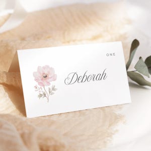May include: A white place card featuring the name "Deborah" in cursive script, with a watercolour illustration of a pink flower. The word "ONE" is printed at the top. The card rests on a beige fabric.