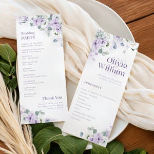 Pode incluir: Dois programas de casamento com desenhos florais em roxo e branco. Os programas apresentam os nomes dos noivos, os membros do casamento e os detalhes da cerimónia. Os programas estão num prato branco com um tecido branco.