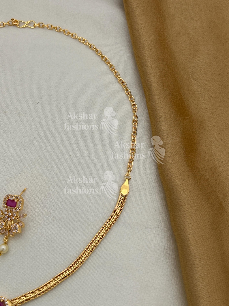May include: A gold-coloured necklace with a woven chain design is shown in close-up. The necklace has a small, square, ruby-coloured stone pendant with a pearl drop. The image also shows a gold-coloured chain and the text "Akshar fashions".