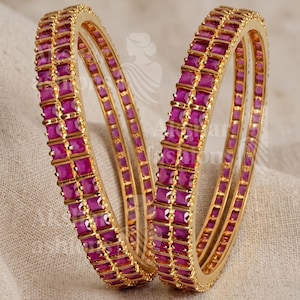 May include: Two gold bangles, each adorned with rows of square, magenta gemstones. The bangles feature a textured gold band and are presented on a neutral surface. The gemstones are uniformly spaced, forming a continuous pattern around the bangles.