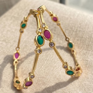 May include: Two gold-toned bangles featuring a series of alternating oval-shaped green and pink stones, interspersed with small, clear, round white stones set along the thin, circular metal bands in a repeating pattern.