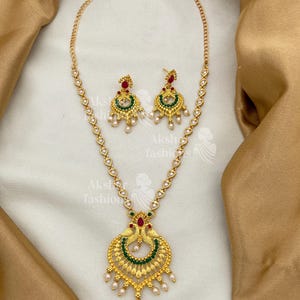 May include: Gold-toned necklace and matching earrings set. The necklace features a pendant with a peacock design, adorned with red, green, and white accents. The earrings mirror the design, with a similar color scheme and pearl-like drops.