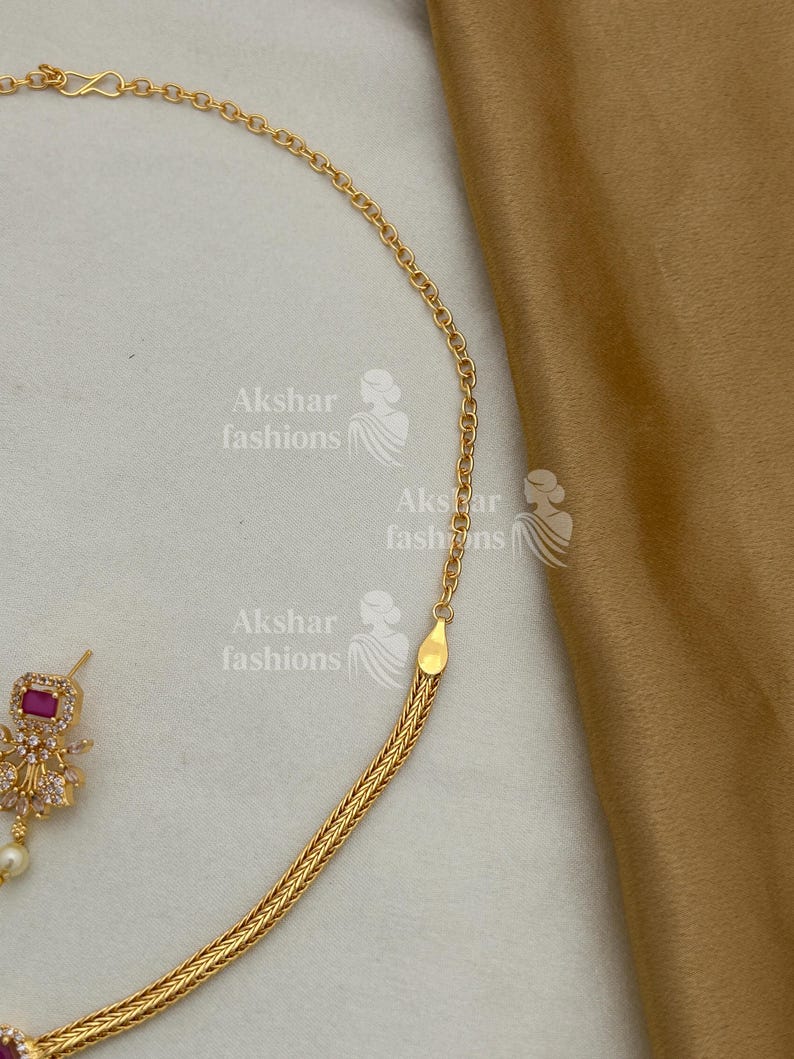 May include: A gold-coloured necklace with a fine chain and a woven section is shown in close-up. The necklace has a small, square, red gemstone pendant with clear stones and a dangling pearl. The background is a light beige fabric.