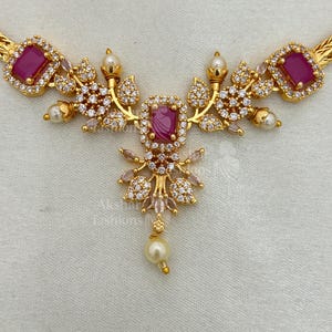 May include: A gold necklace with a central magenta rectangular stone pendant. The necklace is decorated with clear, round accent stones and small, white pearls. The design incorporates floral and leaf-like elements. The necklace is made of gold-toned metal.