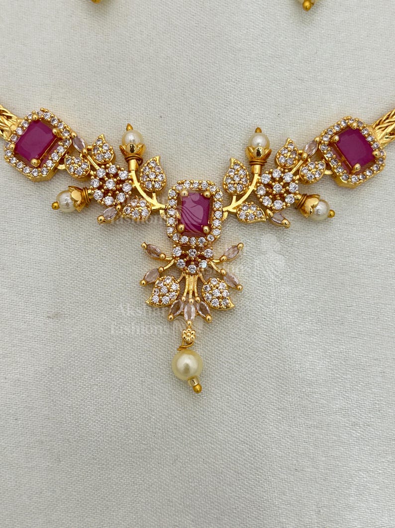 May include: A gold necklace with a central magenta rectangular stone pendant. The necklace is decorated with clear, round accent stones and small, white pearls. The design incorporates floral and leaf-like elements. The necklace is made of gold-toned metal.