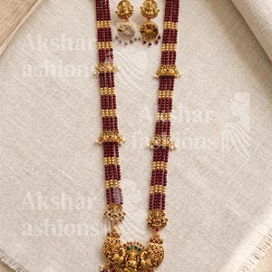May include: An ornate gold-toned necklace and matching earrings set. The necklace features strands of burgundy beads interspersed with gold accents and a detailed pendant with figures and colorful accents. The earrings have a similar design.