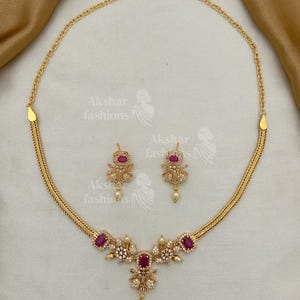 May include: A gold-coloured necklace and earring set. The necklace features a central floral design with a large, rectangular, red gemstone and smaller white gemstones. The earrings mirror the design. The set includes the text "Akshar fashions".