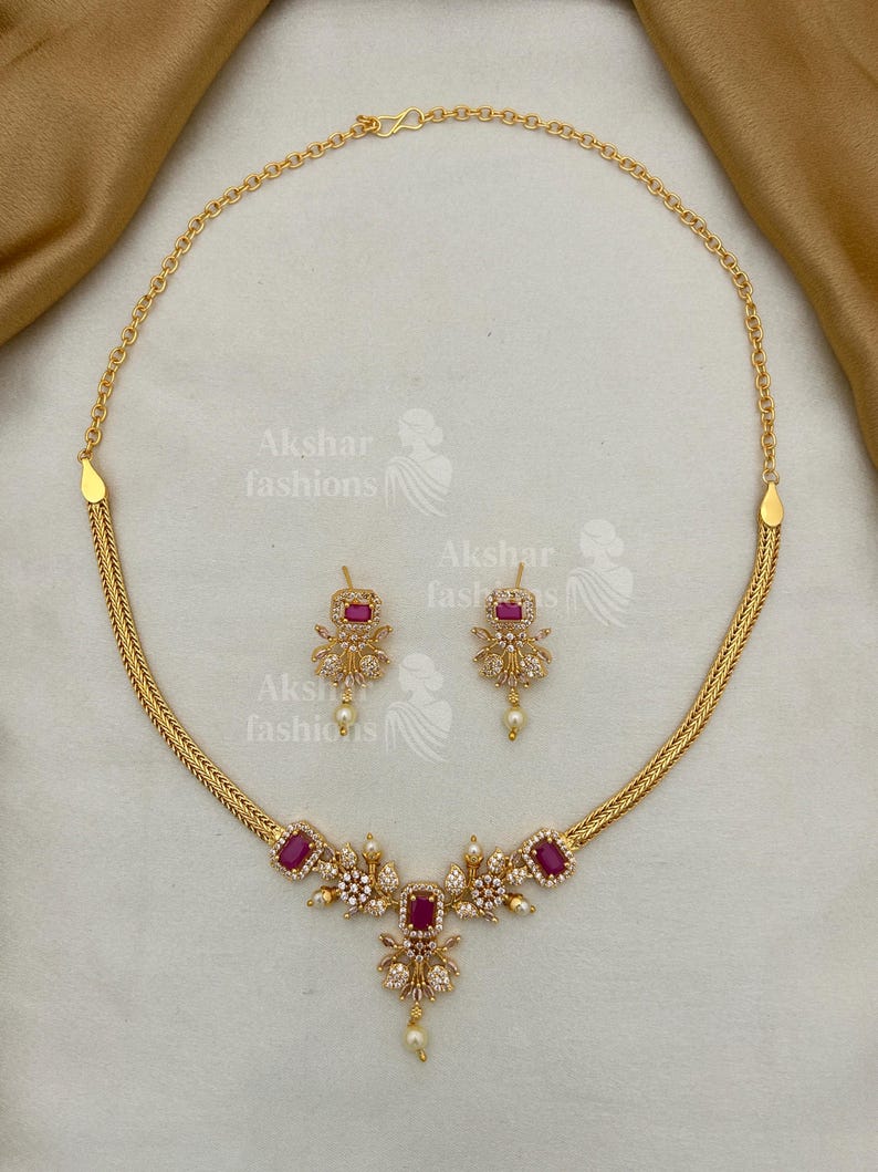 May include: A gold-coloured necklace and earring set. The necklace features a central floral design with a large, rectangular, red gemstone and smaller white gemstones. The earrings mirror the design. The set includes the text "Akshar fashions".