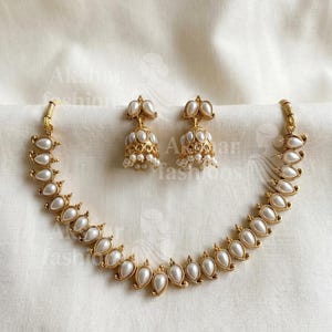 May include: A gold-toned jewelry set featuring a necklace and matching earrings. The necklace and earrings are adorned with pearlescent, teardrop-shaped elements and small, round beads. The set is displayed on a white fabric background.