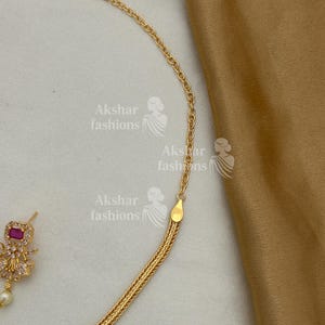 May include: A gold-coloured necklace with a woven chain design is shown. The necklace has a small, square, red gemstone pendant with clear stones and a pearl. The brand name "Akshar fashions" is visible in the image.