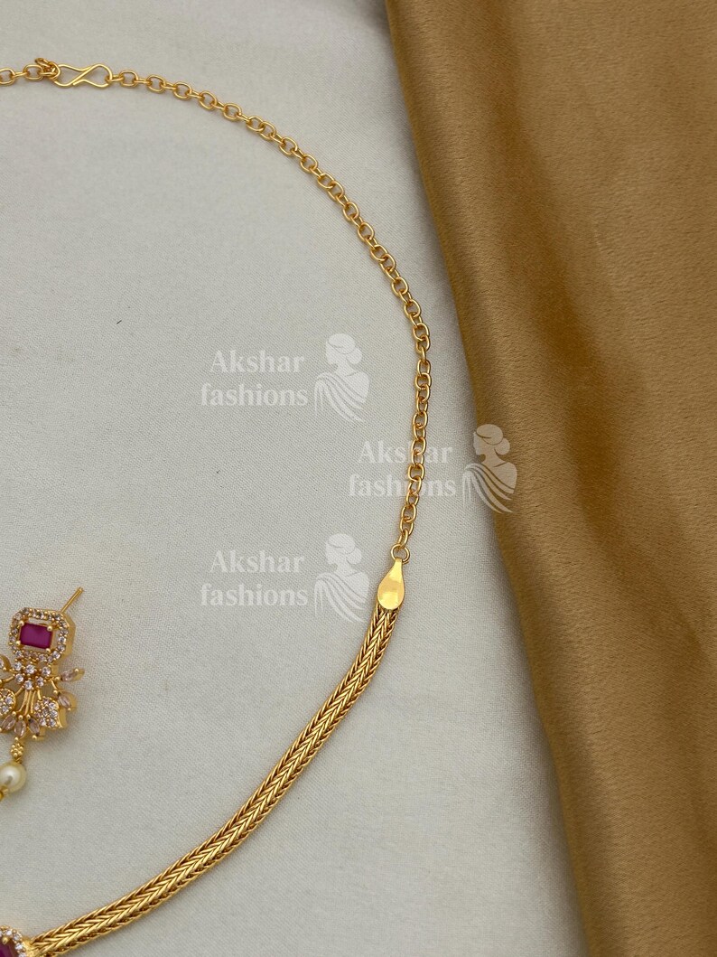 May include: A gold-coloured necklace with a woven chain design is shown. The necklace has a small, square, red gemstone pendant with clear stones and a pearl. The brand name "Akshar fashions" is visible in the image.