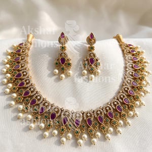May include: A gold-toned necklace and matching earrings set. The necklace and earrings feature teardrop-shaped, deep purple gemstones, small green floral accents, and clear, sparkling stones. White pearls dangle from the necklace and earrings.