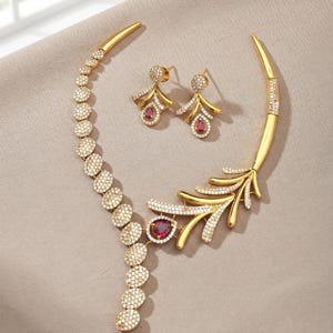May include: A gold-toned necklace and earring set featuring a series of circular links encrusted with small white stones. The necklace includes a prominent leaf-shaped element and a teardrop-shaped red stone. The earrings mirror the teardrop design.