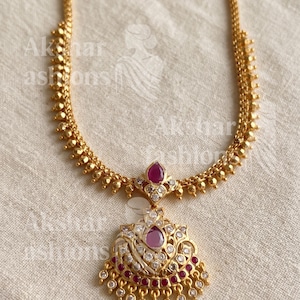 May include: A gold-toned necklace featuring a chain with small spherical beads and a central pendant. The pendant is adorned with a teardrop-shaped pink stone, smaller clear stones, and a row of dangling pink stones at the bottom edge.