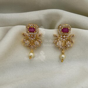 May include: Gold-coloured earrings featuring a rectangular, ruby-red centre stone, encircled by clear crystals. The earrings have a floral design with a dangling pearl. The background is a soft, off-white fabric.