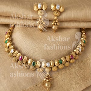 May include: Gold-coloured necklace and earring set. The necklace features oval and spherical elements with colourful gemstones. The earrings have a similar design. The jewellery is displayed on a textured, golden-coloured surface.
