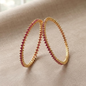 May include: Two circular gold bangles featuring a continuous row of small, square-cut, vibrant pinkish-red stones set along the outer edge. The metal has a polished gold finish, and the design consists of a repeating geometric pattern.
