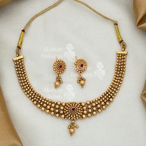 Könnte beinhalten: Ein goldfarbenes Halsketten- und Ohrring-Set. Die Halskette hat ein Perlendesign mit einem zentralen Blumenanhänger und einem roten Edelstein. Die passenden Ohrringe haben ein ähnliches Blumendesign mit einem roten Edelstein und einer hängenden Perle.