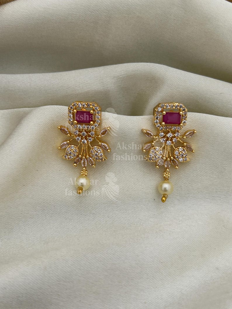 May include: Gold-toned earrings with a rectangular, ruby-coloured centre stone. The earrings have a floral design with clear, sparkling accents and a dangling pearl. The earrings are displayed on a cream-coloured fabric.