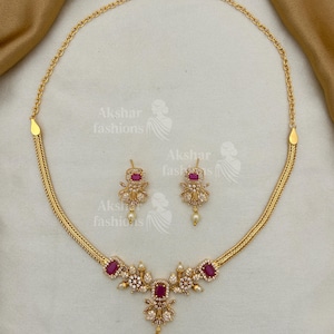 May include: A gold-toned necklace and earring set. The necklace features a central floral design with a large, rectangular, red gemstone and smaller white gemstones. The earrings mirror the design. The set includes the text "Akshar fashions".